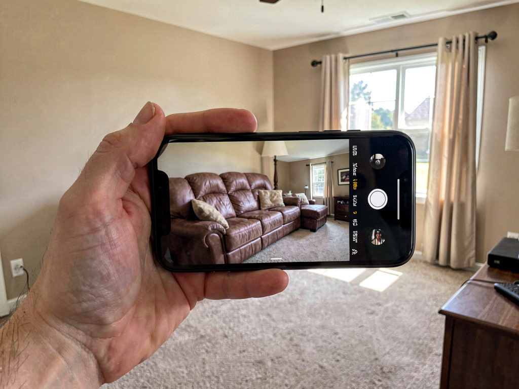 A person standing in a doorway holding up a smartphone to photograph their living room, with natural light from a window