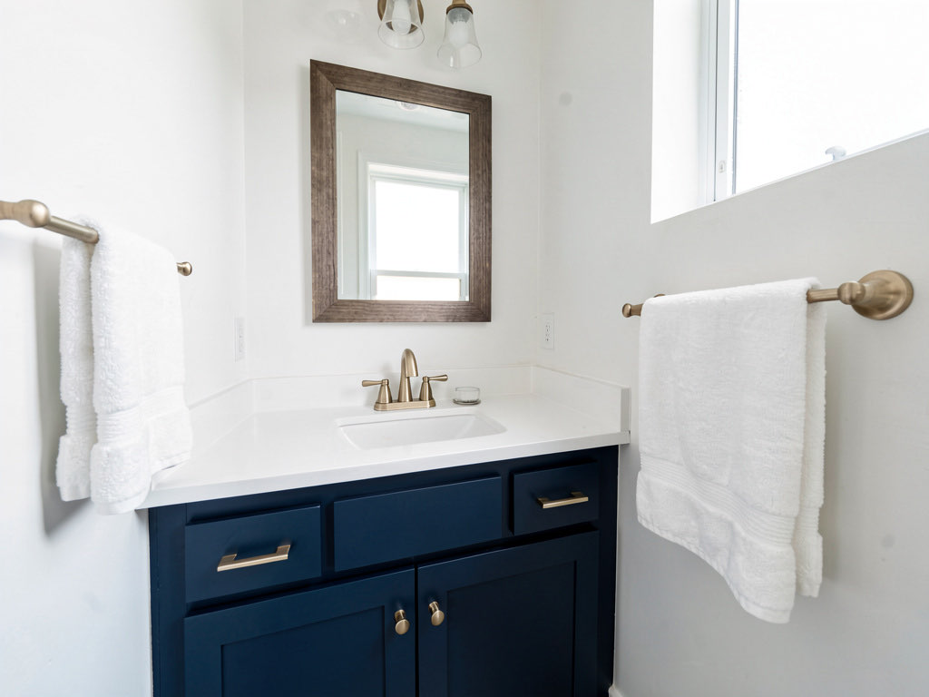 A small bathroom freshly updated with white walls, a navy blue painted vanity, brushed gold hardware and faucet, a new framed mirror, and white towels on a new towel bar