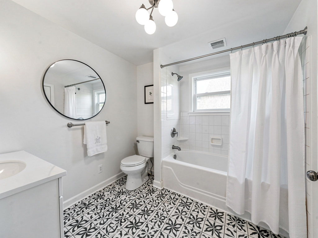 A bathroom with new black and white peel-and-stick floor tile, fresh white paint, an updated three-globe light fixture, a new round mirror, and a new shower curtain