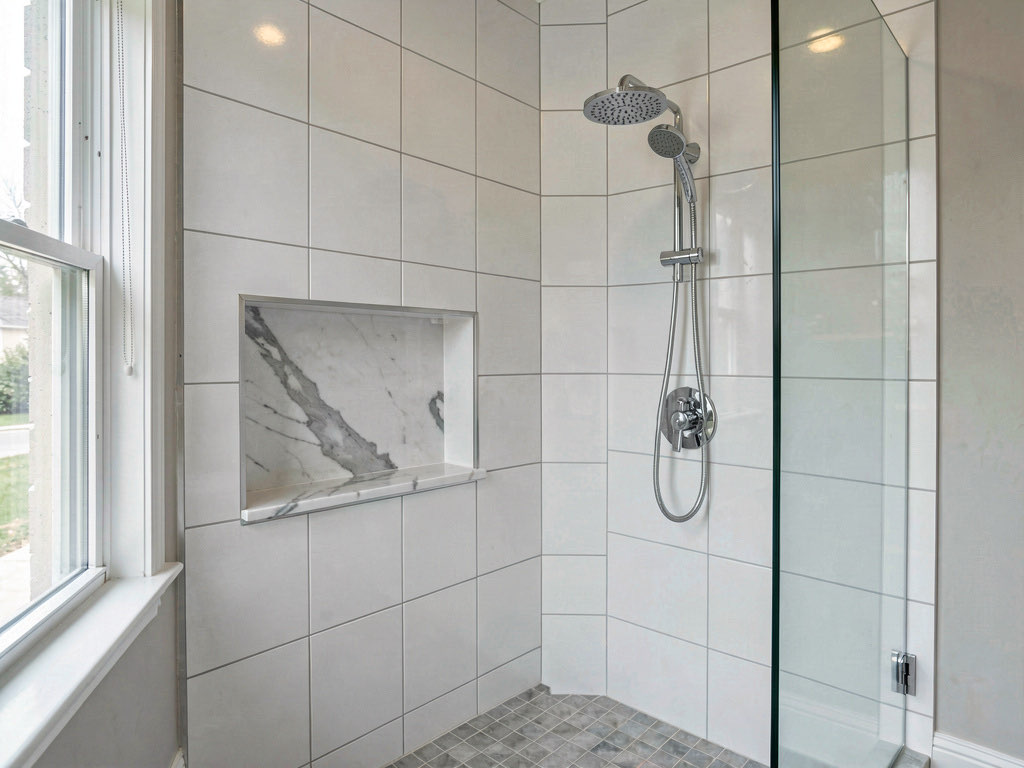 A freshly retiled shower with large format white porcelain tiles, a built-in niche with marble shelf, chrome rain showerhead, and frameless glass door