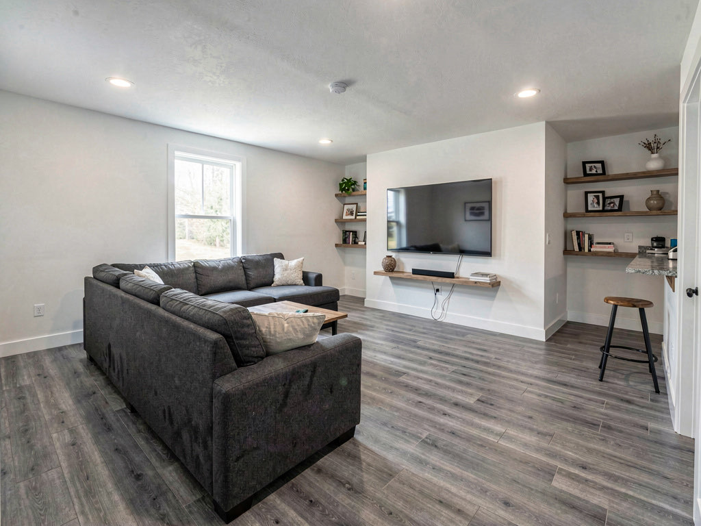 A finished basement with gray luxury vinyl flooring, white walls, recessed lighting, charcoal sectional sofa, mounted TV, and a small bar area