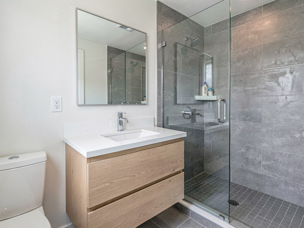 A mid-range bathroom remodel with floating light wood vanity, white quartz top, gray tile shower, frameless glass door, and chrome fixtures
